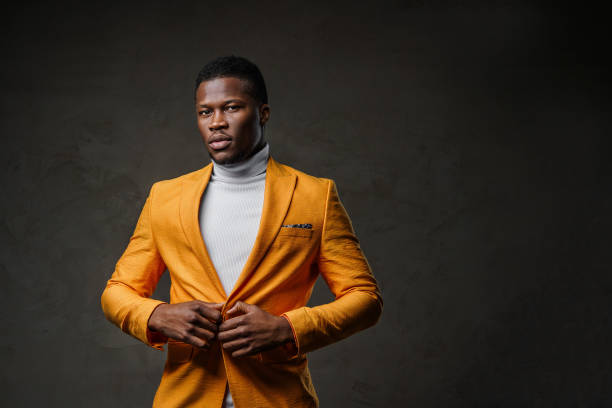 A stylish dark-skinned man posing while buttoning his yellow jacket against a dark backdrop